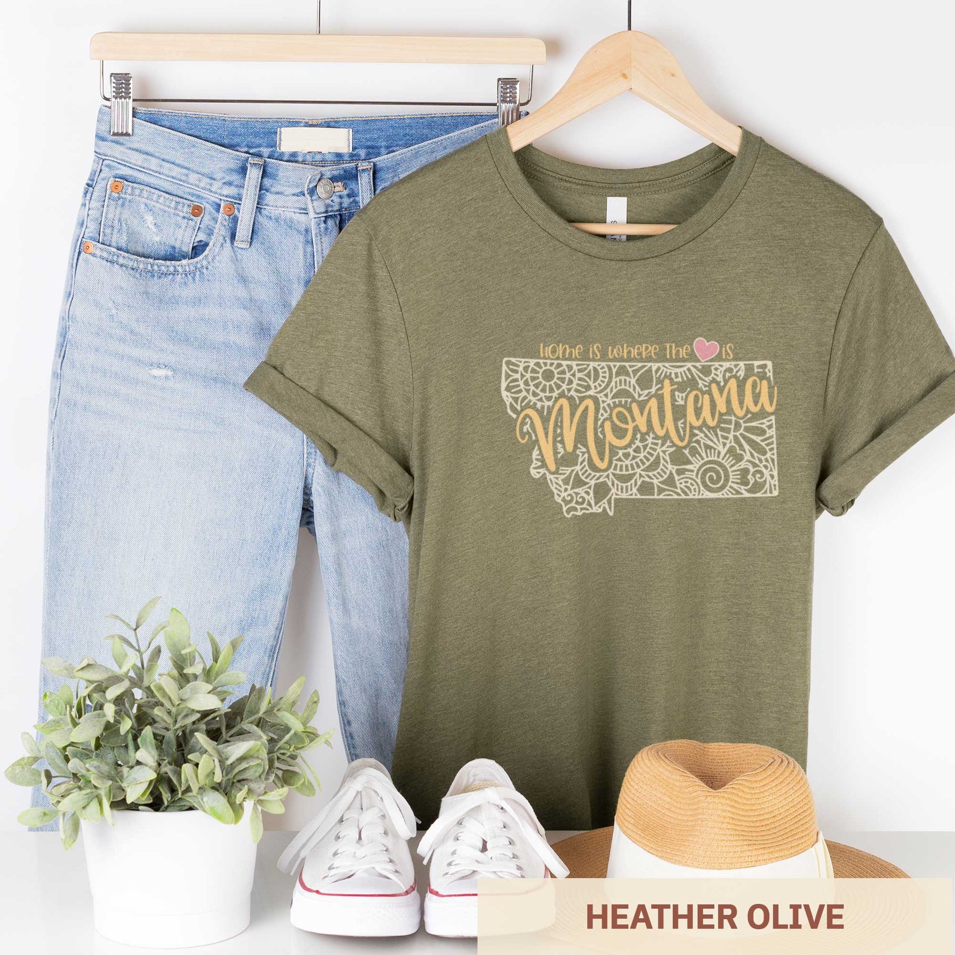A hanging heather olive Bella Canvas t-shirt featuring a mandala in the shape of Montana with the words home is where the heart is.