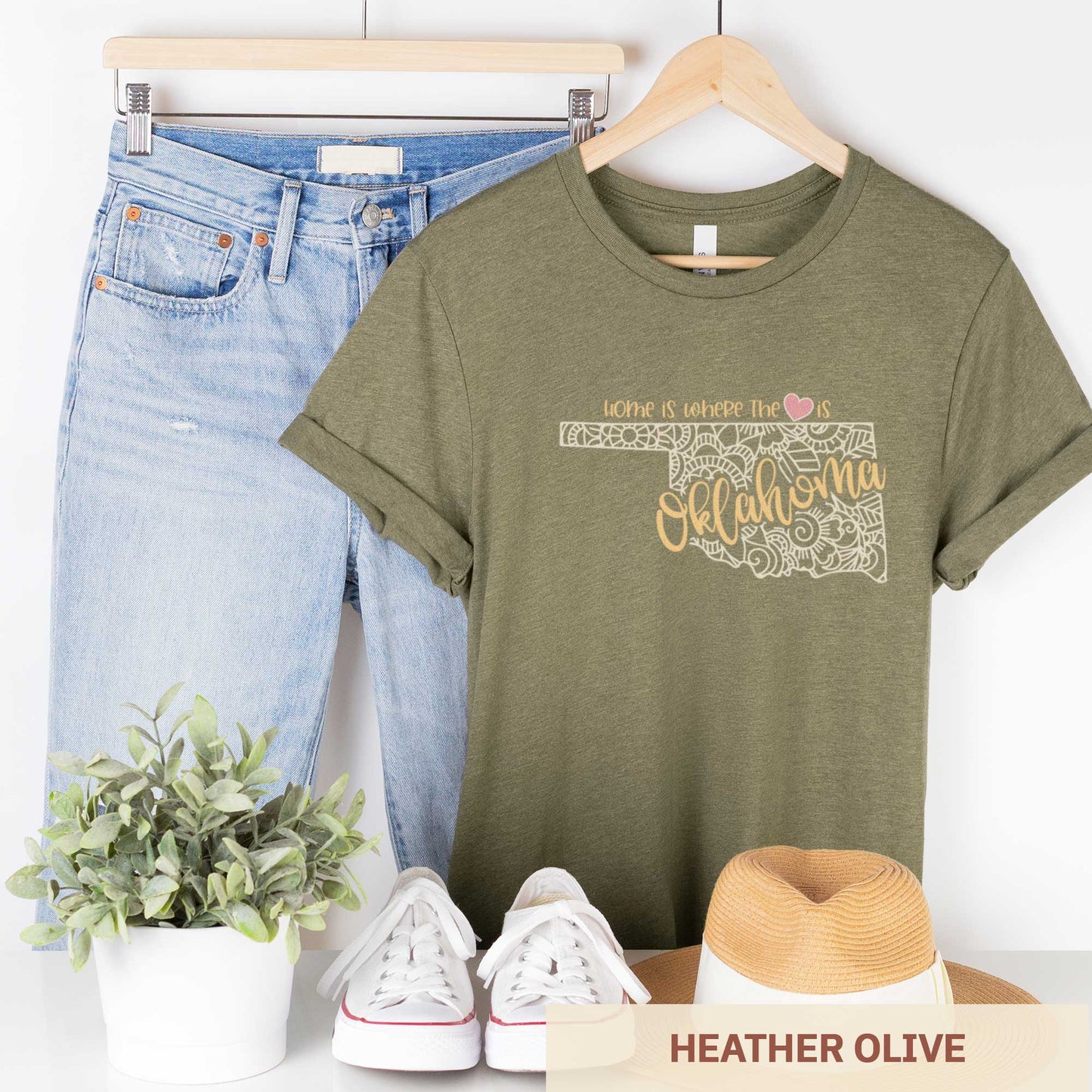 A hanging heather olive Bella Canvas t-shirt featuring a mandala in the shape of Oklahoma with the words home is where the heart is.