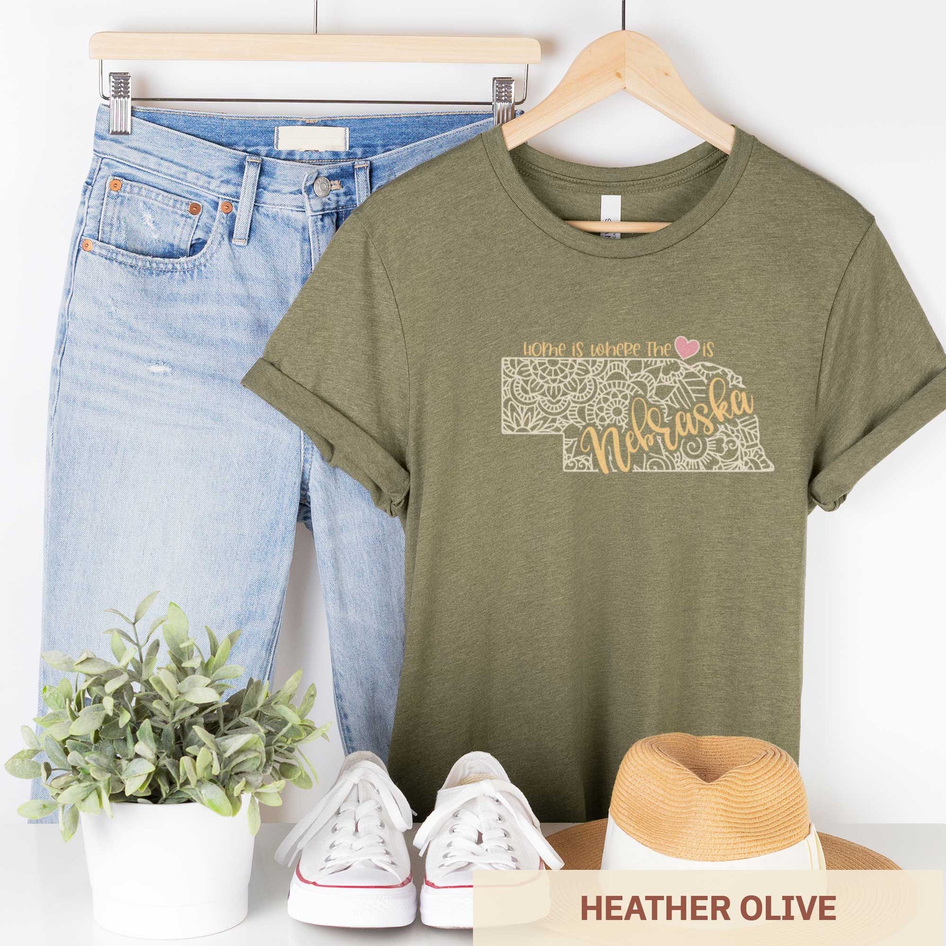 A hanging heather olive Bella Canvas t-shirt featuring a mandala in the shape of Nebraska with the words home is where the heart is.