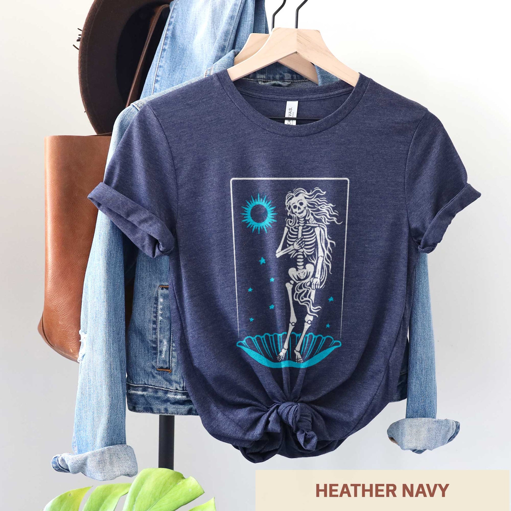 A hanging heather navy Bella Canvas t-shirt that features a skeleton in a pose similar to Botticelli's Birth of Venus painting.