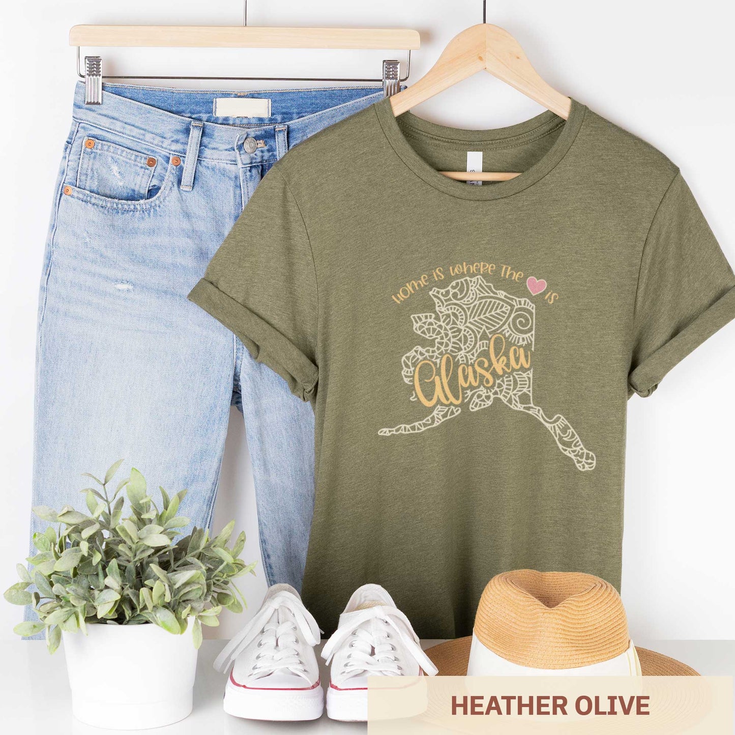 A hanging heather olive Bella Canvas t-shirt featuring a mandala in the shape of Alaska with the words home is where the heart is.