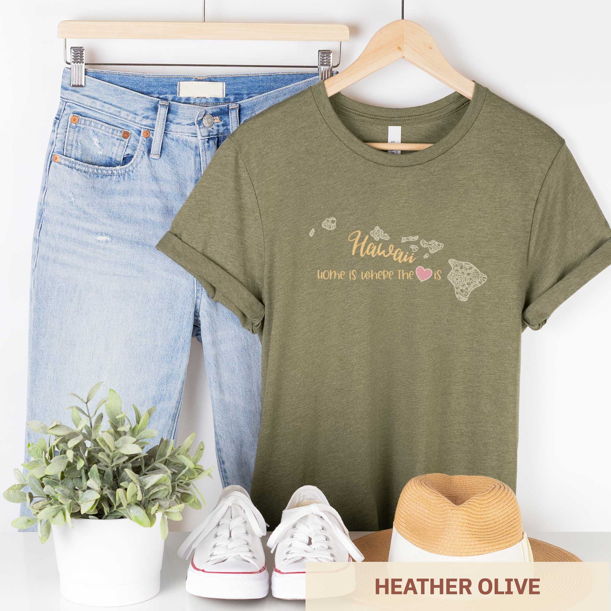 A hanging heather olive Bella Canvas t-shirt featuring a mandala in the shape of Hawaii with the words home is where the heart is.