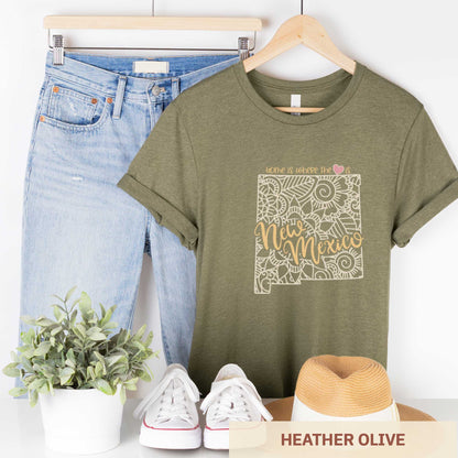 A hanging heather olive Bella Canvas t-shirt featuring a mandala in the shape of New Mexico with the words home is where the heart is.