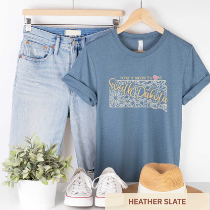 A hanging heather slate Bella Canvas t-shirt featuring a mandala in the shape of South Dakota with the words home is where the heart is.