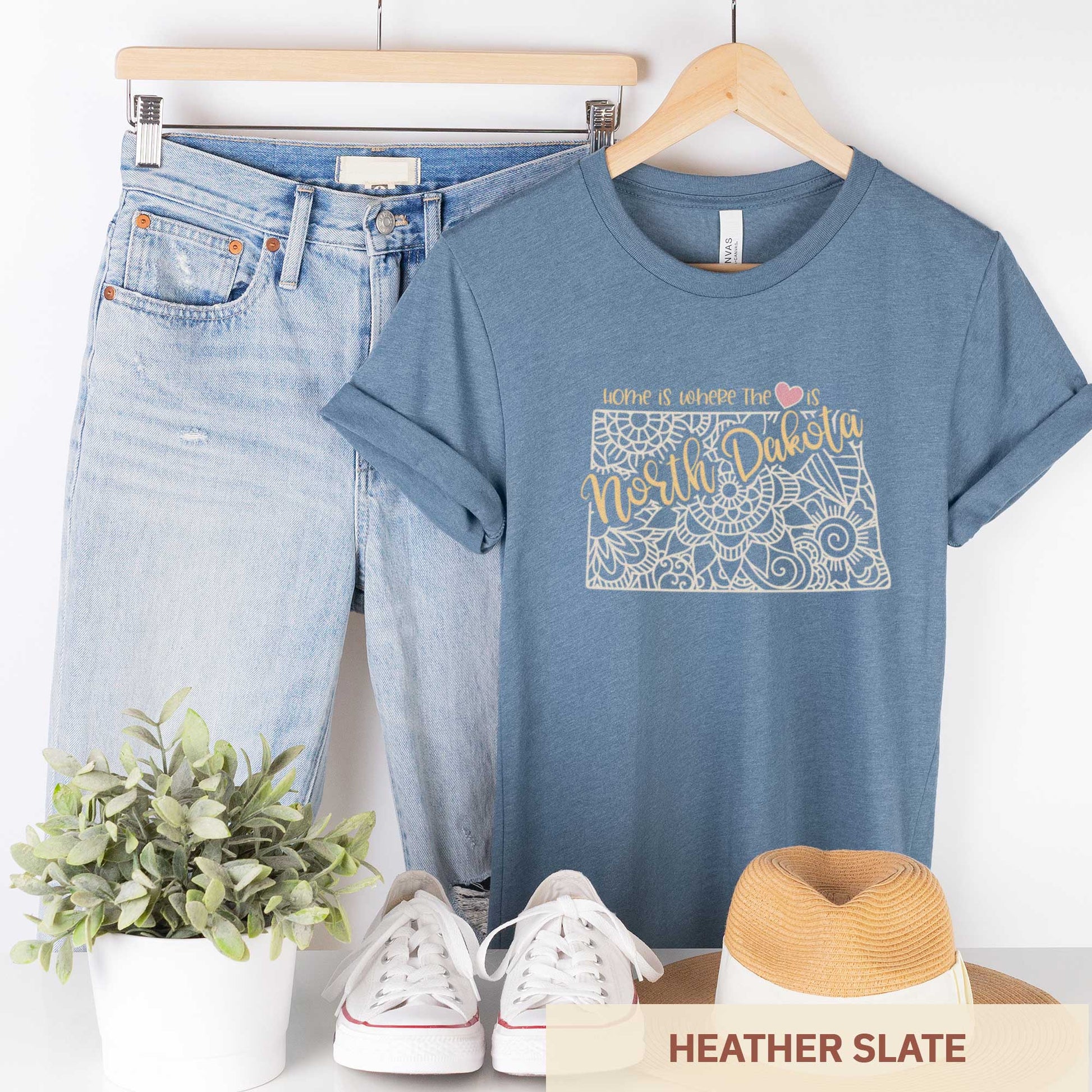A hanging heather slate Bella Canvas t-shirt featuring a mandala in the shape of North Dakota with the words home is where the heart is.
