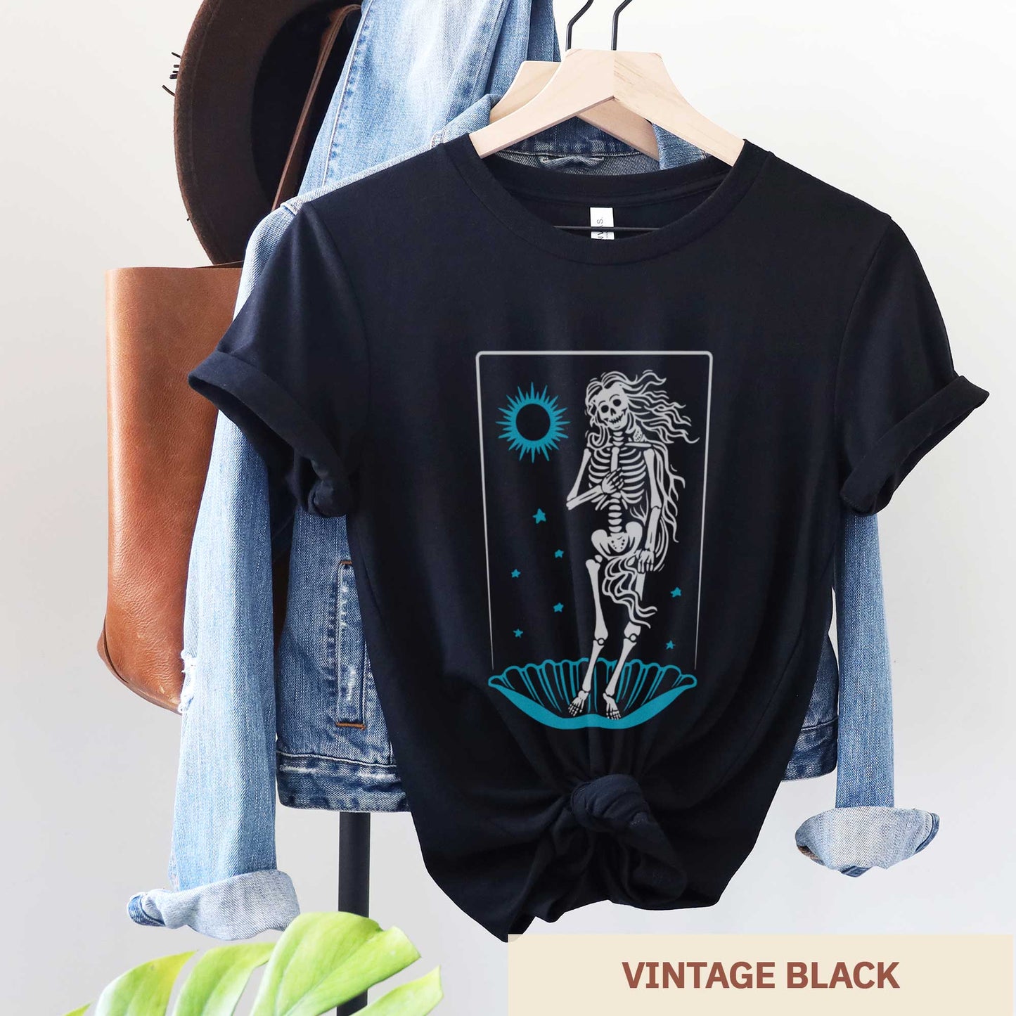 A hanging vintage black Bella Canvas t-shirt that features a skeleton in a pose similar to Botticelli's Birth of Venus painting.
