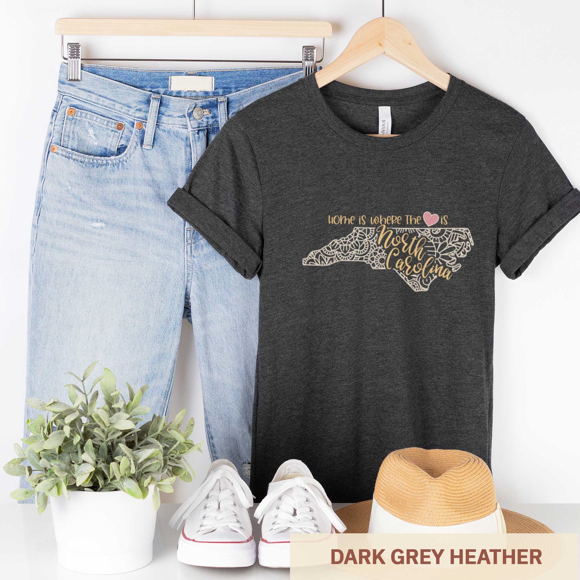 A hanging dark grey heather Bella Canvas t-shirt featuring a mandala in the shape of North Carolina with the words home is where the heart is.
