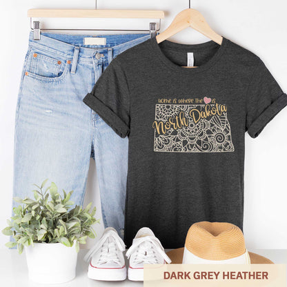 A hanging dark grey heather Bella Canvas t-shirt featuring a mandala in the shape of North Dakota with the words home is where the heart is.