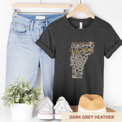 A hanging dark grey heather Bella Canvas t-shirt featuring a mandala in the shape of Vermont with the words home is where the heart is.