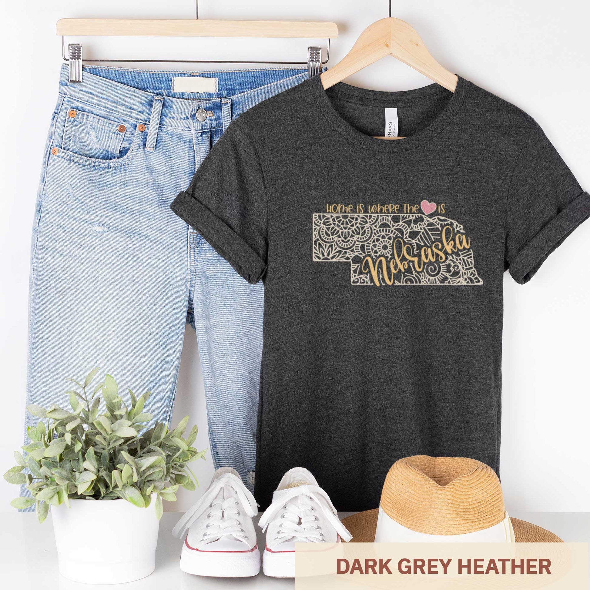 A hanging dark grey heather Bella Canvas t-shirt featuring a mandala in the shape of Nebraska with the words home is where the heart is.