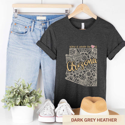 A hanging dark grey heather Bella Canvas t-shirt featuring a mandala in the shape of Arizona with the words home is where the heart is.