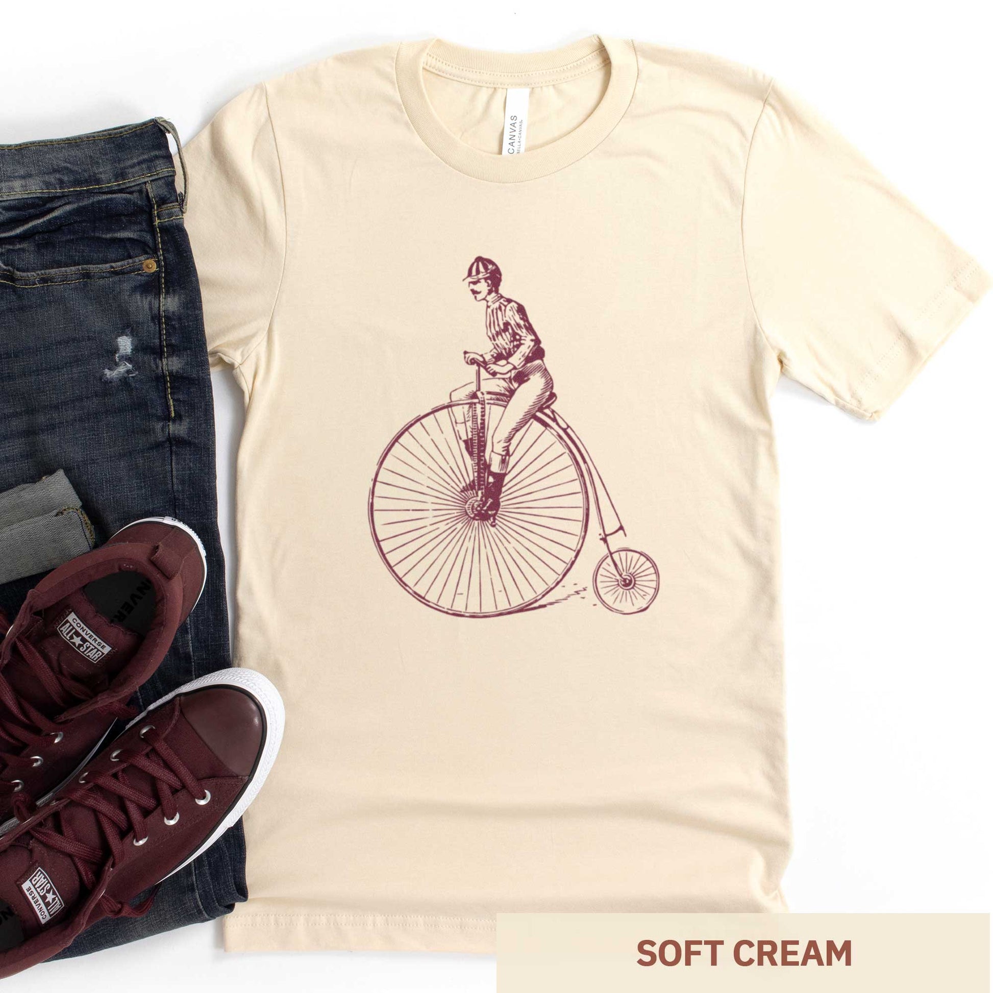 A soft cream Bella Canvas t-shirt featuring a Victorian man riding a penny farthing bicycle.