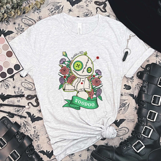 An ash Bella Canvas t-shirt surrounded by goth boots and makeup and featuring a cartoon voodoo doll and flowers.