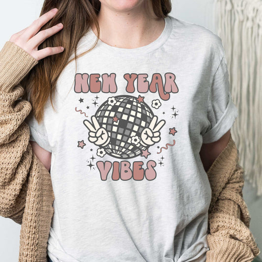 A woman wearing an ash Bella Canvas t-shirt featuring a retro disco ball holding up hands in a peace sign that says New Year vibes.