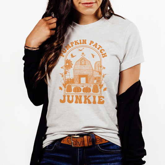 A woman wearing an ash Bella Canvas t-shirt featuring a farm with pumpkins that says pumpkin patch junkie.