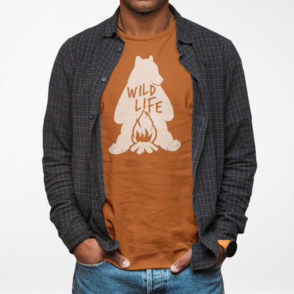 A man wearing an autumn burnt orange Bella Canvas t-shirt featuring a silhouette of a grizzly bear sitting in front of a campfire with the words wild life.