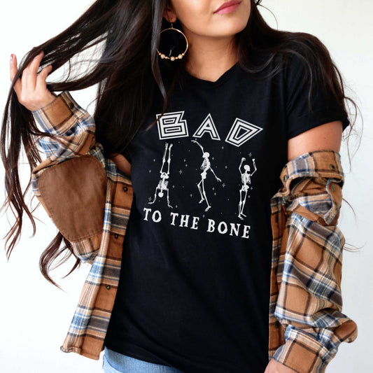 A woman wearing a black Bella Canvas t-shirt that says bad to the bone surrounded by skeletons.