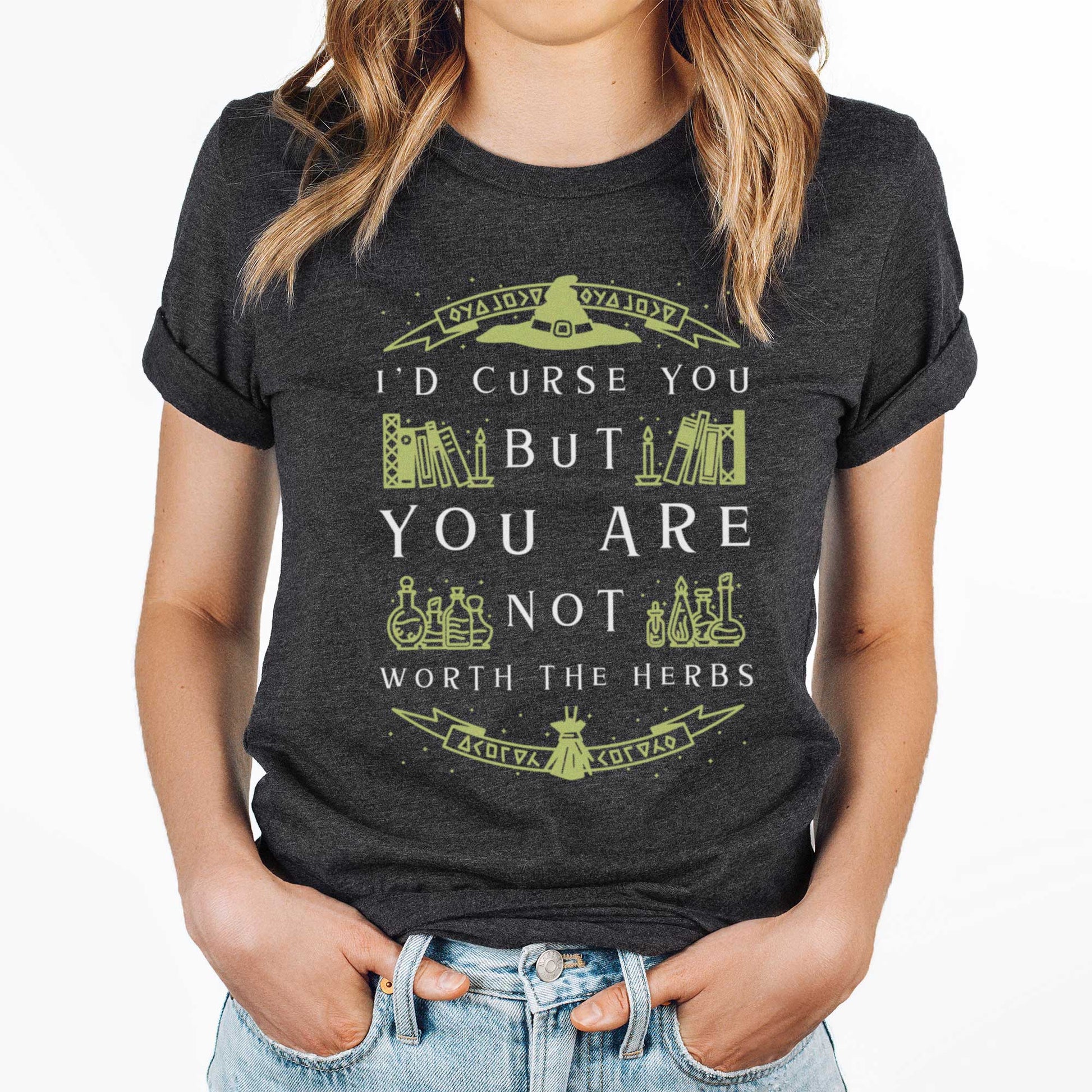 A woman wearing a dark grey heather Bella Canvas t-shirt with a witch's hat, books and potions and the words I'd curse you but you are not worth the herbs.