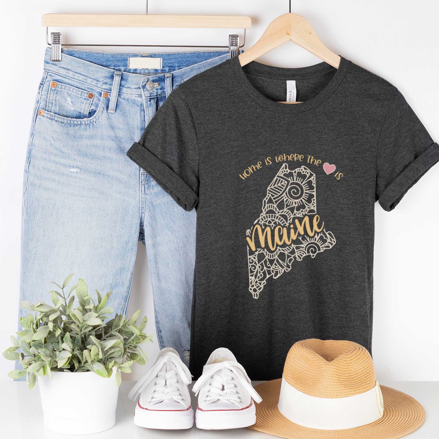 A hanging dark grey heather Bella Canvas t-shirt featuring a mandala in the shape of Maine with the words home is where the heart is.