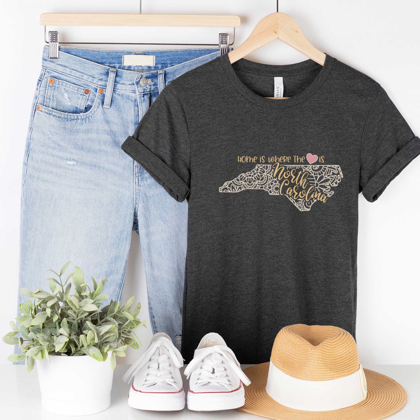 A hanging dark grey heather Bella Canvas t-shirt featuring a mandala in the shape of North Carolina with the words home is where the heart is.