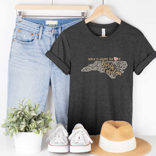 A hanging dark grey heather Bella Canvas t-shirt featuring a mandala in the shape of North Carolina with the words home is where the heart is.