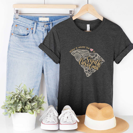 A hanging dark grey heather Bella Canvas t-shirt featuring a mandala in the shape of South Carolina with the words home is where the heart is.