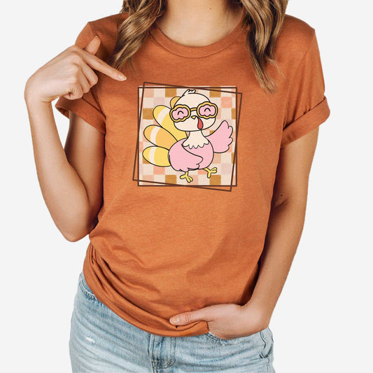A woman wearing a heather autumn Bella Canvas t-shirt featuring a cute cartoon turkey against a fall checkered background.