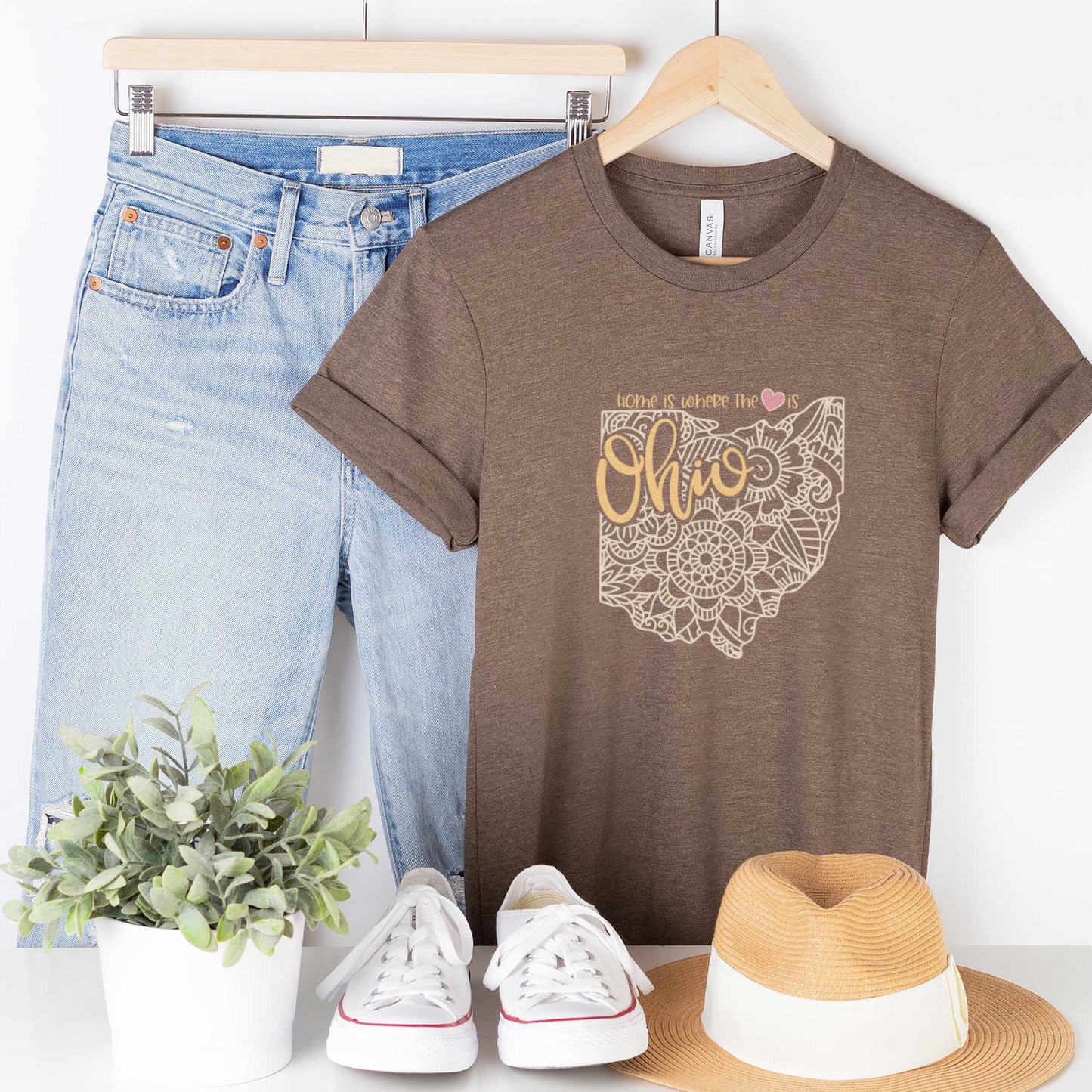 A hanging heather brown Bella Canvas t-shirt featuring a mandala in the shape of Ohio with the words home is where the heart is.