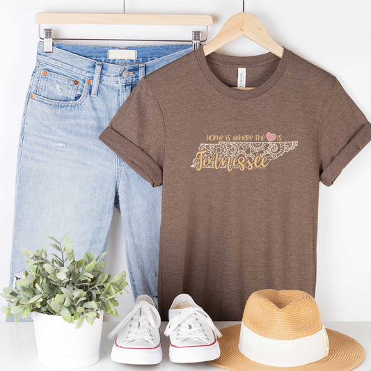 A hanging heather brown Bella Canvas t-shirt featuring a mandala in the shape of Tennessee with the words home is where the heart is.