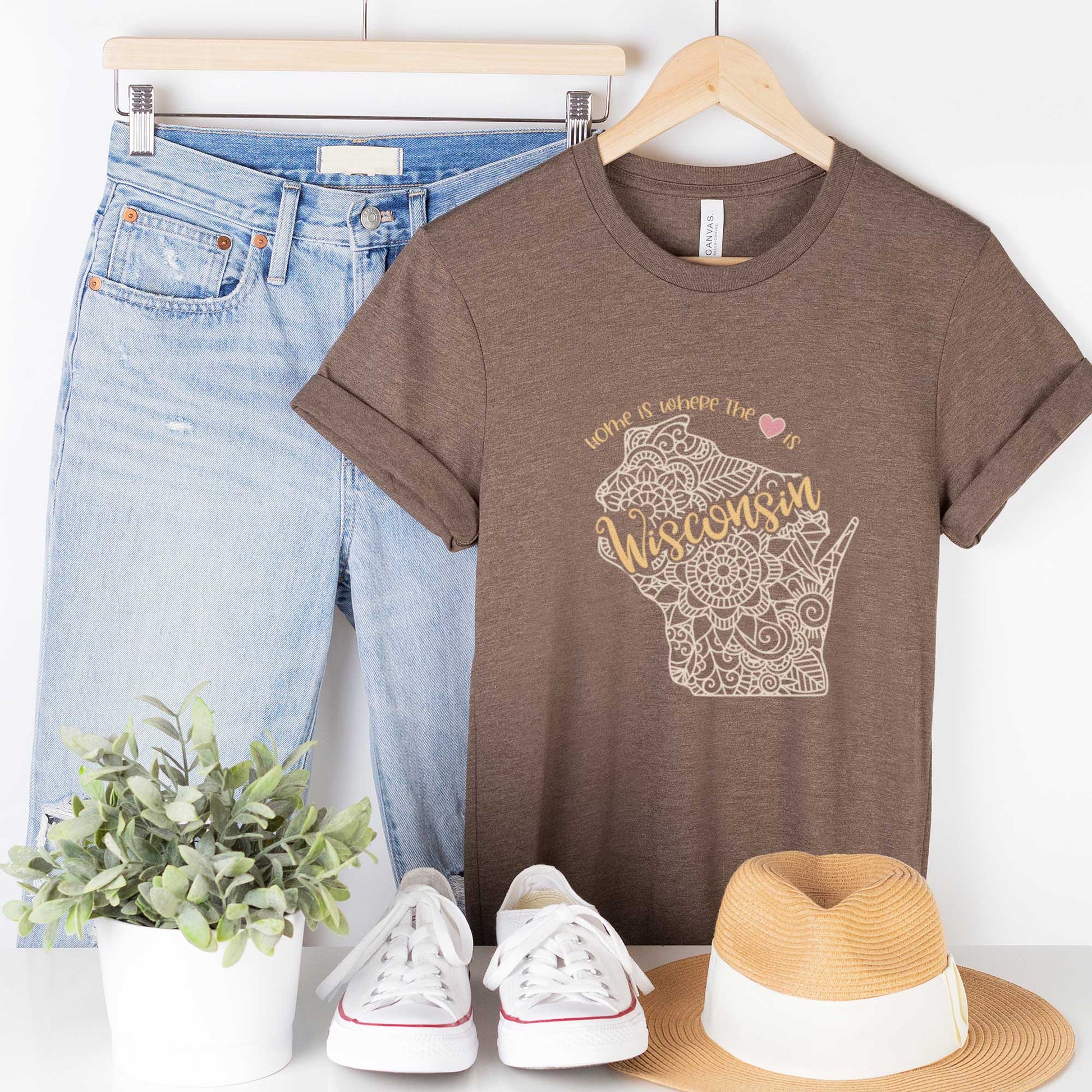 A hanging heather brown Bella Canvas t-shirt featuring a mandala in the shape of Wisconsin with the words home is where the heart is.