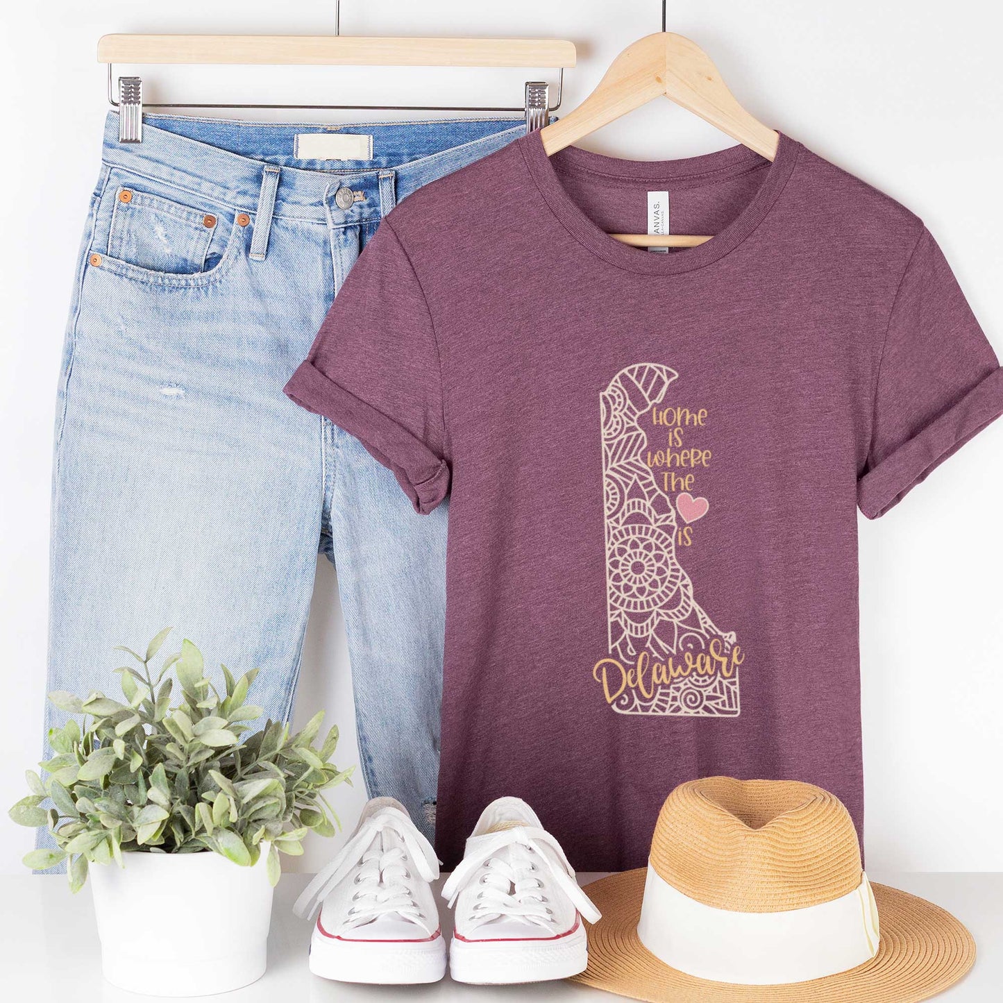 A hanging heather maroon Bella Canvas t-shirt featuring a mandala in the shape of Delaware with the words home is where the heart is.