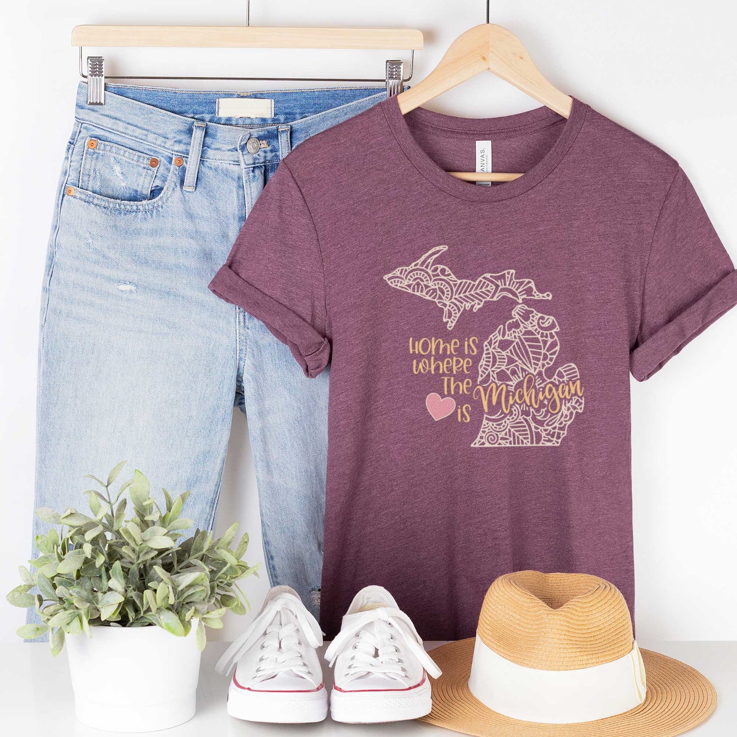 A hanging heather maroon Bella Canvas t-shirt featuring a mandala in the shape of Michigan with the words home is where the heart is.
