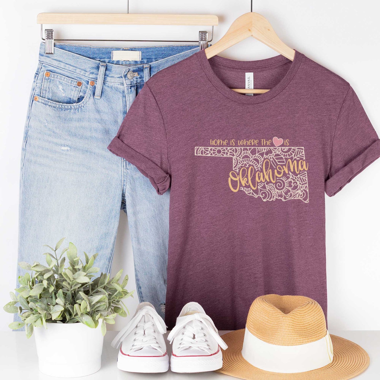 A hanging heather maroon Bella Canvas t-shirt featuring a mandala in the shape of Oklahoma with the words home is where the heart is.