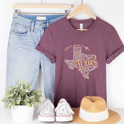 A hanging heather maroon Bella Canvas t-shirt featuring a mandala in the shape of Texas with the words home is where the heart is.