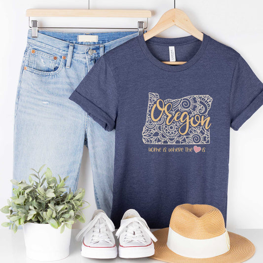 A hanging heather navy Bella Canvas t-shirt featuring a mandala in the shape of Oklahoma with the words home is where the heart is.