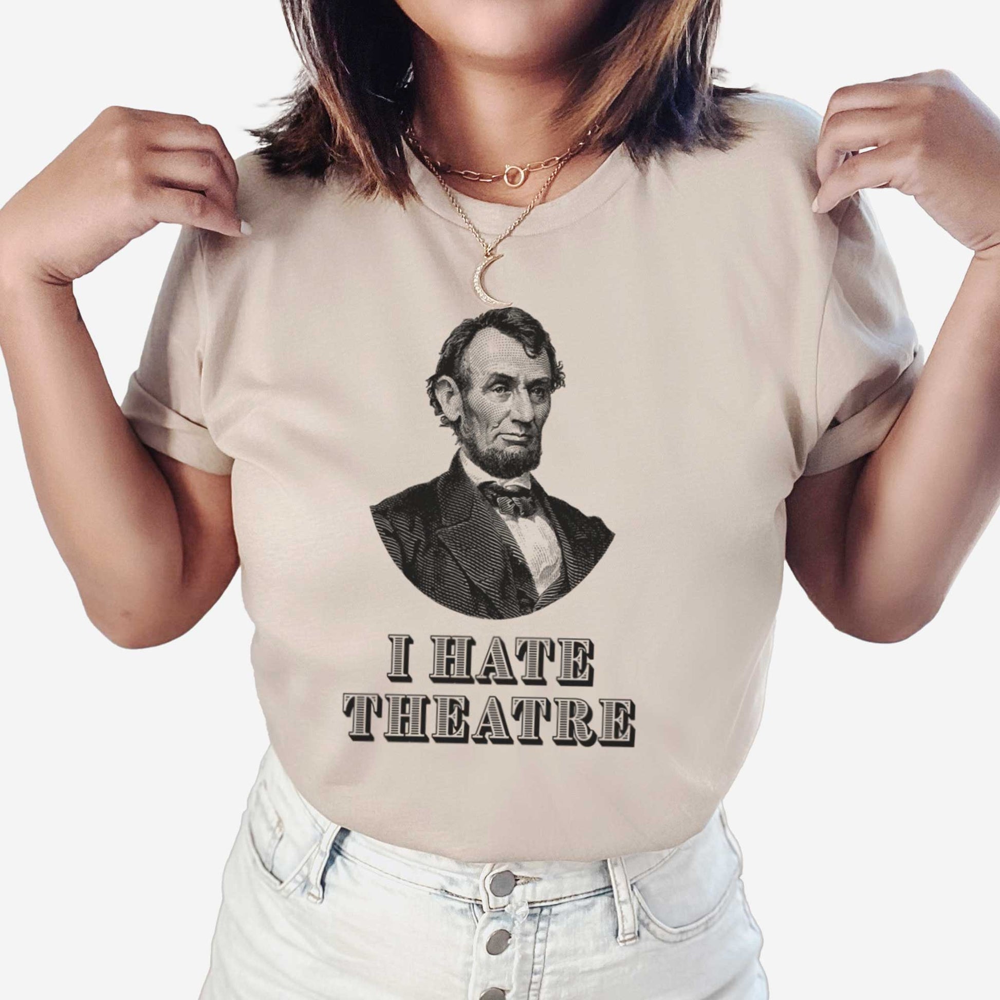 A woman wearing a heather tan Bella Canvas t-shirt featuring a vintage illustration of Abraham Lincoln with the words I hate theatre below it.