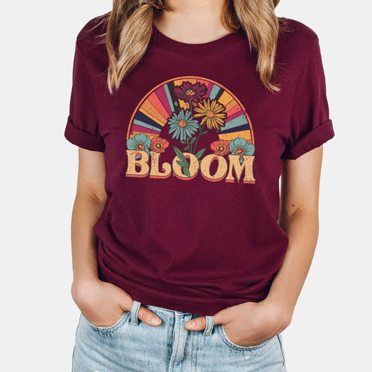 A woman wearing a maroon red Bella Canvas t-shirt featuring a retro styled illustration of flowers and a multi-colored sunray with the words bloom.