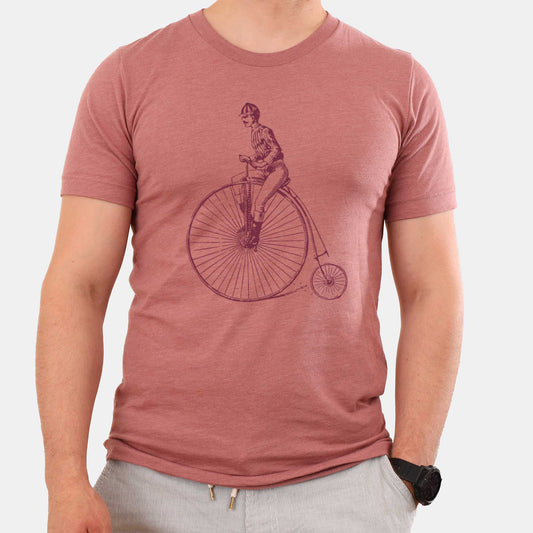 A man wearing a heather mauve Bella Canvas t-shirt featuring a Victorian man riding a penny farthing bicycle.