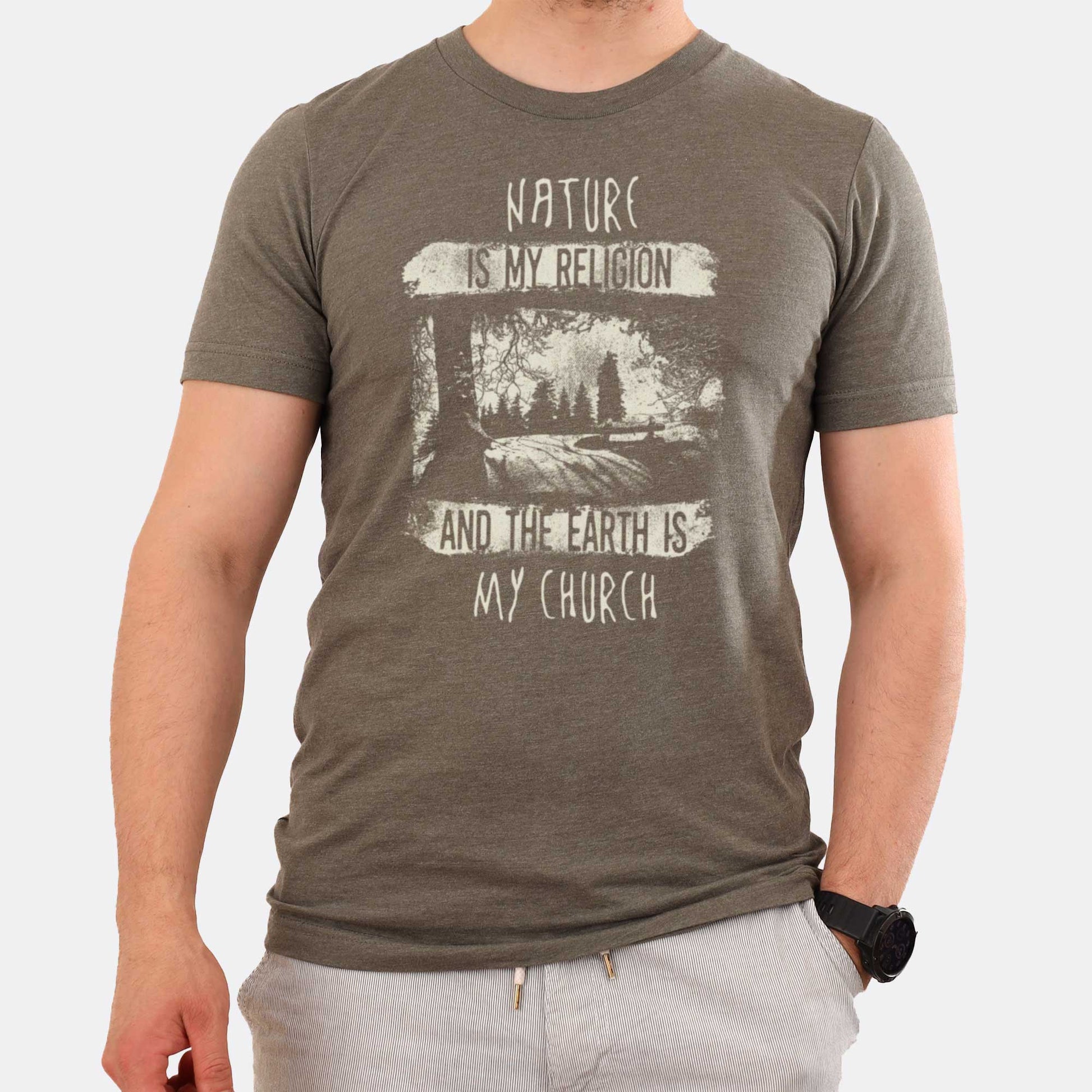A man wearing a heather military green Bella Canvas t-shirt featuring fields and a forest with the words nature is my religion and the earth is my church.