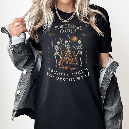 A woman wearing a black Bella Canvas t-shirt with dancing skeletons in front of a fire and an ouija board layout.