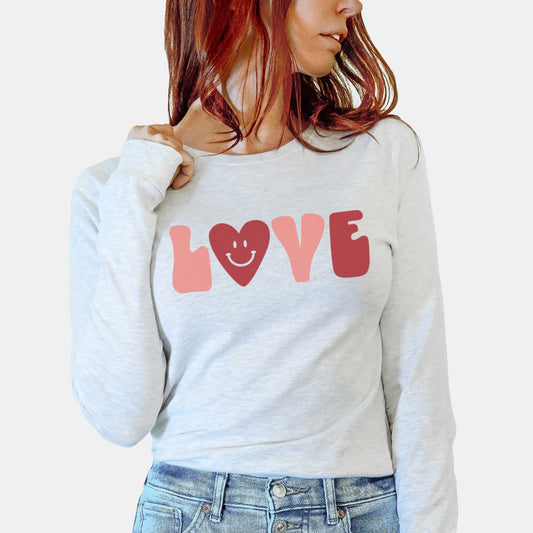 A woman wearing an ash Bella Canvas long sleeve t-shirt featuring the words love in a retro styled font where the "o" has been replaced by a heart smiley face.