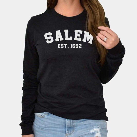 A woman wearing a black Bella Canvas long sleeve t-shirt that says Salem Est. 1692..