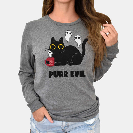 A woman wearing a grey Bella Canvas long sleeve t-shirt featuring a cartoon black cat drinking a satanic cup of coffee with ghosts around it and the words purr evil.