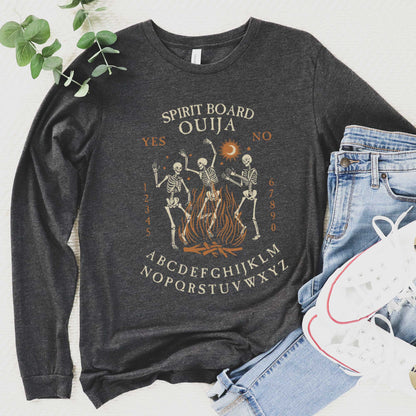 A dark grey heather Bella Canvas long sleeved t-shirt with dancing skeletons in front of a fire and an ouija board layout.