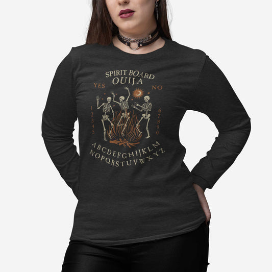 A woman wearing a dark grey heather Bella Canvas long sleeve t-shirt with dancing skeletons in front of a fire and an ouija board layout.