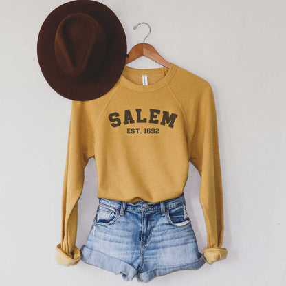A hanging mustard yellow Bella Canvas sweatshirt that says Salem Est. 1692