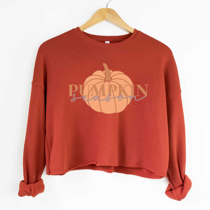 A hanging brick red Bella Canvas cropped sweatshirt featuring a light orange pumpkin with the words pumpkin season over it.