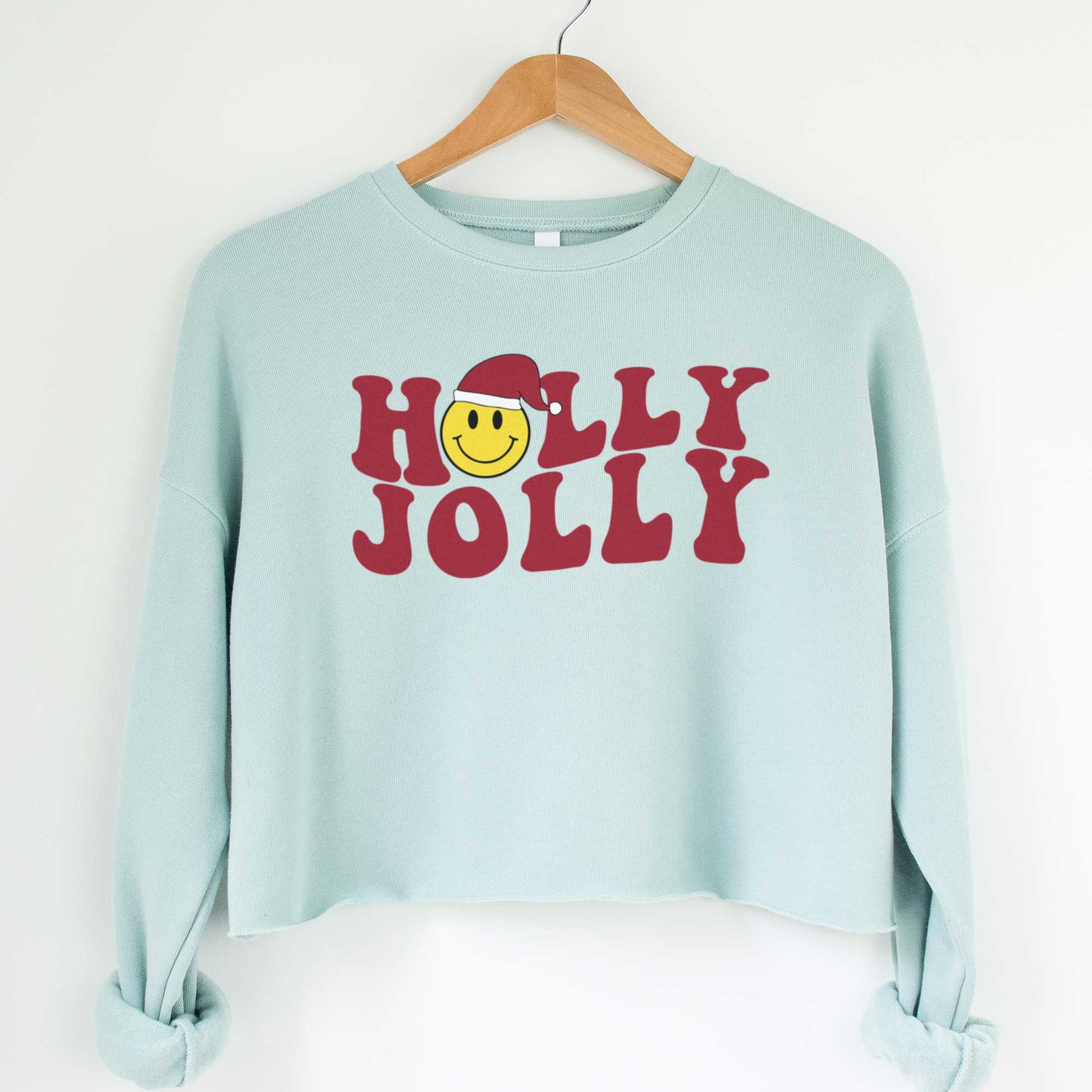 A hanging dusty blue cropped Bella Canvas sweatshirt featuring a yellow smiley face with a santa hat among the words holly jolly.