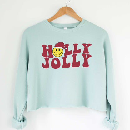 A hanging dusty blue cropped Bella Canvas sweatshirt featuring a yellow smiley face with a santa hat among the words holly jolly.