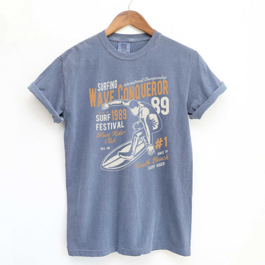 A hanging blue jean Comfort Colors t-shirt featuring a man riding a surfboard and the words wave conqueror